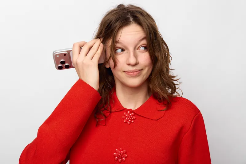 Young woman in red sweater holds smartphone near ear, looking sideways with curious expression. Isolated on white background. Concept of communication, technology, and casual lifestyle.