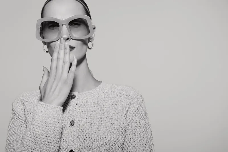 Stylish Caucasian woman with fair skin wearing oversized sunglasses and textured sweater covers mouth with hand in studio. Concept of fashion and elegance.
