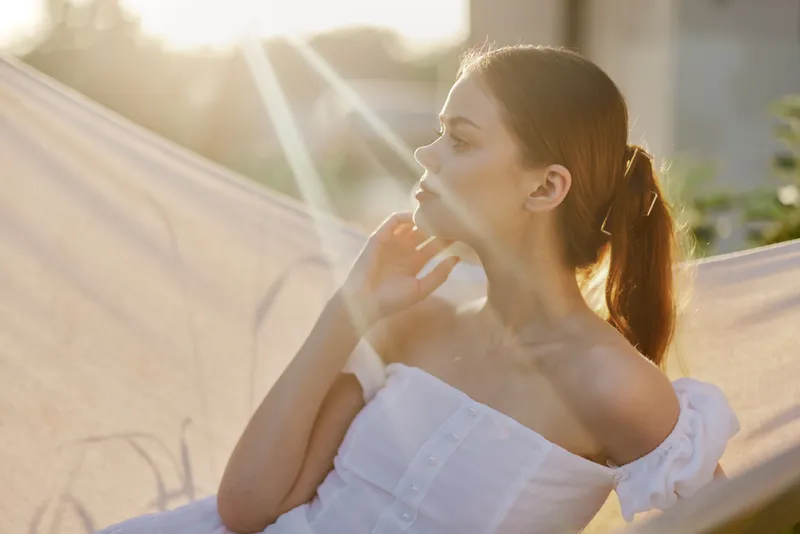 Young woman in white dress enjoying sunset, relaxing in hammock, with soft lighting creating a serene atmosphere, capturing feelings of tranquility and peace