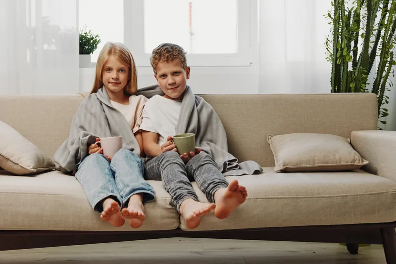 Two children wrapped in blankets sit barefoot on a sofa drinking from cups in a cozy living room with natural light and plants.