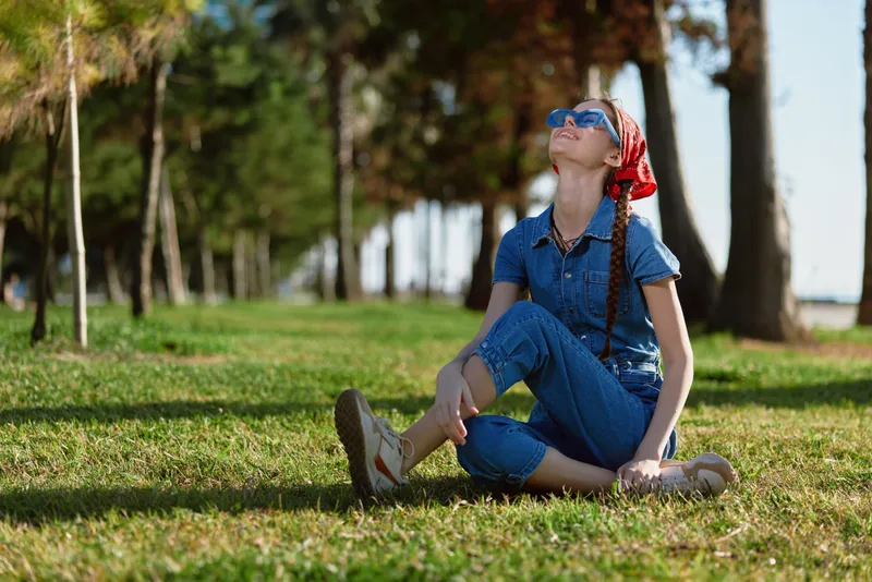 Lifestyle woman in modern boho-western denim street style sitting on grass outdoors wearing sunglasses and a red bandana with film color tones and natural background