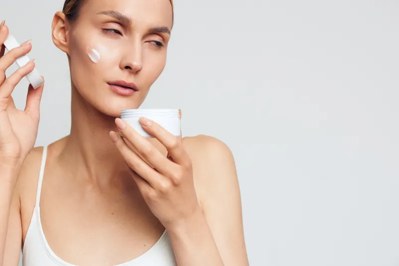 A young woman applies face cream in a bright indoor studio, calm expression conveys a focused skincare moment during a clean routine