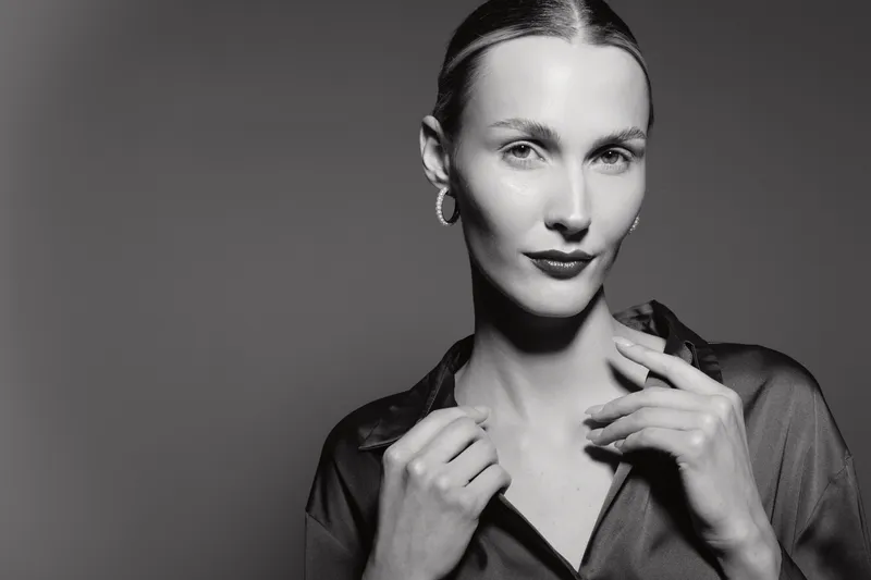 Portrait of a white female model with light skin and slicked hair wearing earrings and a sheer blouse in monochrome studio lighting.