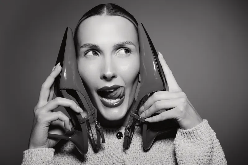 Happy Caucasian woman with light skin holding high heel shoes near face with playful expression in black and white studio portrait. Concept of fashion, fun, and lifestyle.