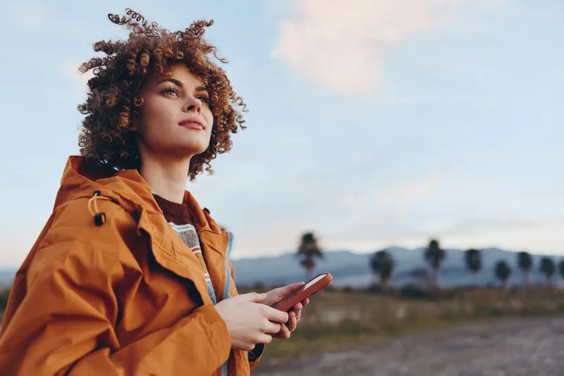 Woman lifestyle outdoors, curly hair, orange jacket portrait, holding smartphone while looking up, sunlit sunset sky, calm relaxed mood, natural field background