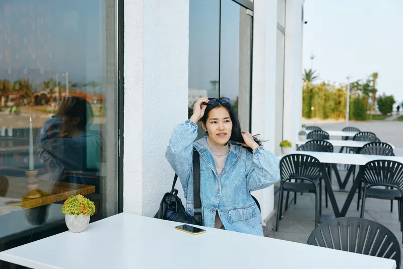 Woman at an outdoor cafe adjusting sunglasses beside a glass wall, denim jacket and casual style in a sunlit urban setting, capturing a relaxed daytime mood and contemporary fashion moment