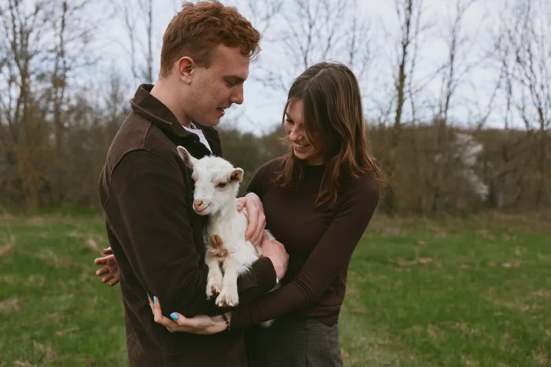 Couple outdoors with a young goat, sharing a gentle moment in a grassy field. Warm, natural lighting highlights affection, connection, and rural lifestyle. Tender bonding with animals.