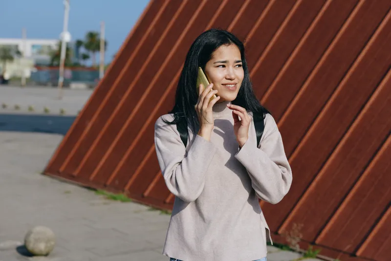Woman on cellphone outdoors beside a rusted wall, wearing a light knit sweater, casual urban scene with sunlit pavement, showing communication, mobility and everyday chatter in a friendly daytime