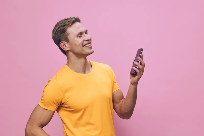 smiling man holding smartphone in orange t-shirt look side positive emotion isolated pink background digital detox concept communication technology lifestyle