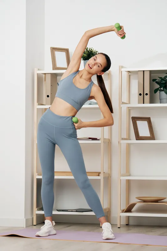 Woman in gray athletic wear performing overhead dumbbell stretch in a bright home gym, showcasing fitness, workout routine, and balance during a focused exercise session.
