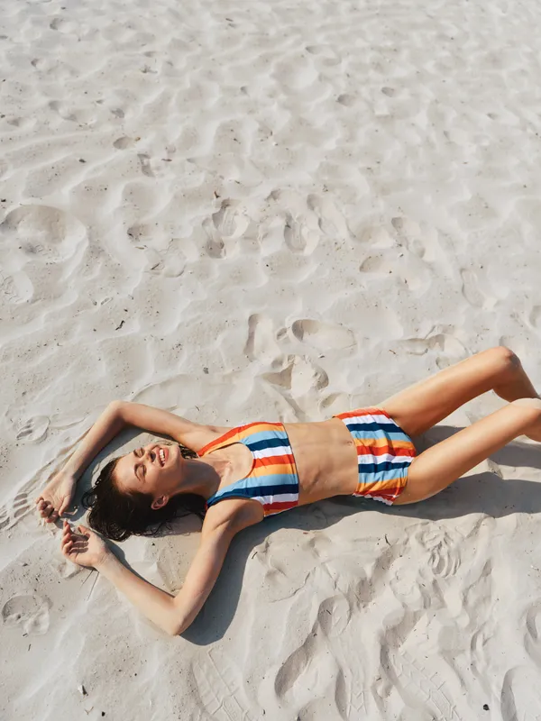 A woman lies sunbathing on white sand, wearing a striped bikini and relaxed pose, enjoying warm sunlight and a peaceful coastal moment outdoors.
