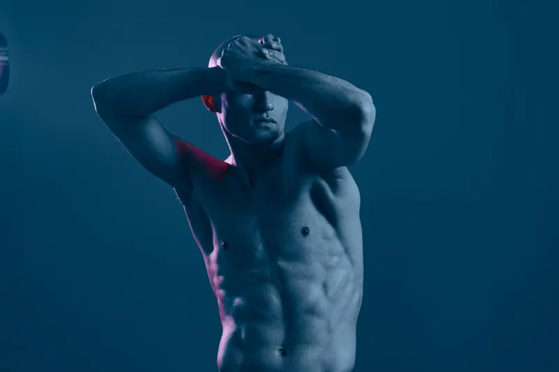 Strong man posing dramatically with his arms crossed, showcasing muscular physique and expressing emotion in a moody blue light. This image captures intensity and power.