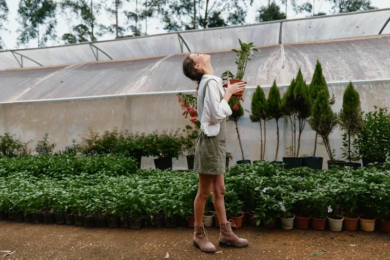 Young woman in casual work attire smiling joyfully in a lush greenhouse, surrounded by vibrant green plants and decorative pots, conveying a sense of nature and happiness