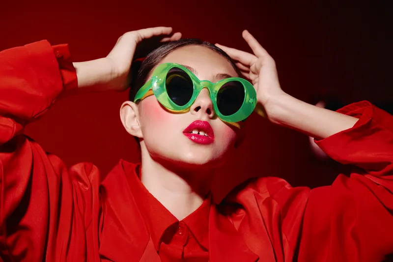 Young woman wearing vibrant green sunglasses and bright red lipstick poses confidently with hands on head. She wears a red outfit against a dark red background. Concept of beauty, fashion, and color