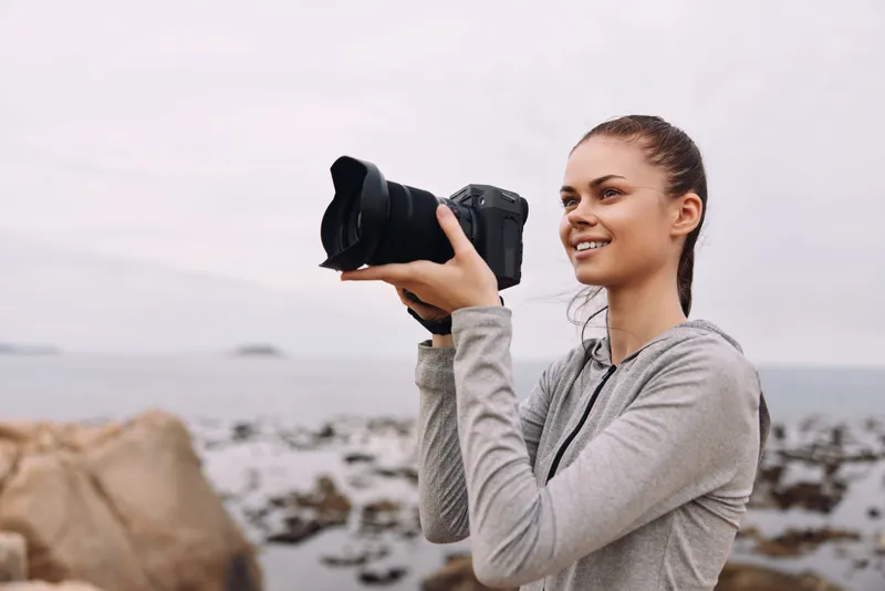 Young woman with a camera capturing nature s beauty by the coast, wearing a gray hoodie, smiling with confidence in a serene outdoor setting