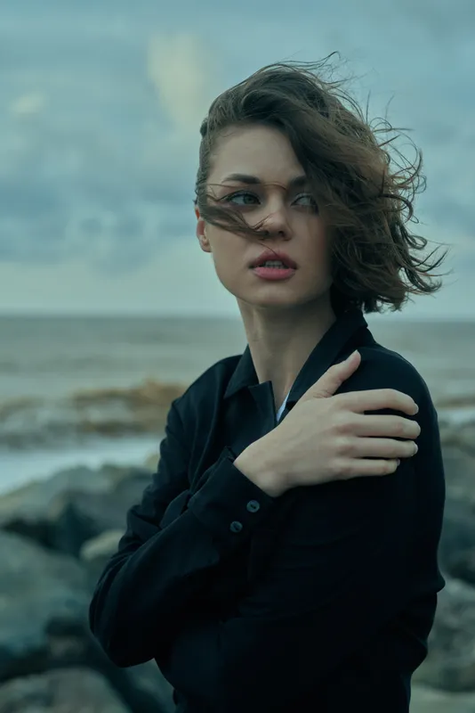 Woman on a windy coastline wears a dark shirt, hair blown by sea breeze, contemplative mood, moody lighting, rugged rocks and vast ocean in the background, atmospheric fashion and editorial scene