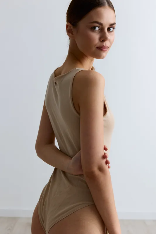 Young woman posing in beige bodysuit with neutral expression against plain light background, showcasing natural beauty and minimalist style in soft indoor lighting.