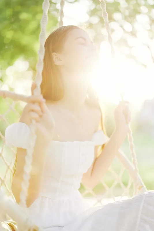 A serene young woman in a white dress joyfully swinging in the sunlight, conveying happiness and tranquility amidst a lush green background