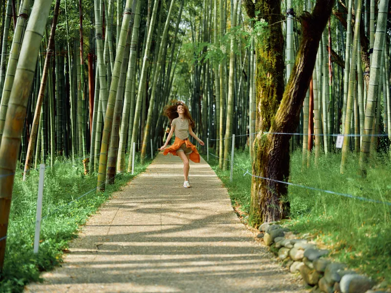 Girl jumping on a path through a bamboo forest wearing a hat and orange skirt, running carefree along a shaded walkway with sunlight, motion and joyful summer outdoor energy.