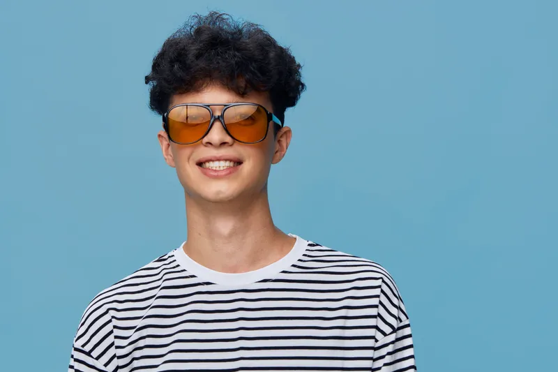 young man wearing striped shirt and orange sunglasses smiling cheerfully against blue background, casual style with curly hair and bright mood