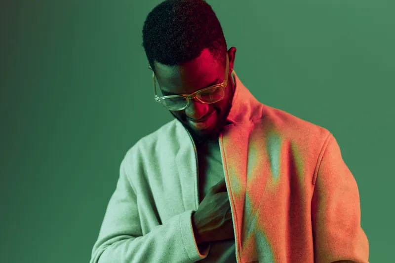 Stylish African American man in a light beige coat with glasses, smiling against a vibrant green background, exuding confidence and modern fashion trends