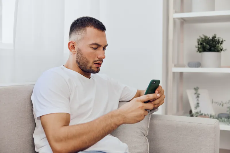 Focused man using smartphone while sitting on a cozy sofa, dressed casually in a white t-shirt. Perfect blend of relaxation and technology in modern living space.