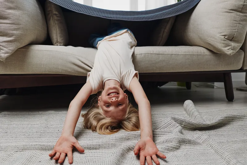 Happy child hanging upside down from sofa with arms stretched on carpet, smiling and playful in cozy living room. Concept for childhood joy, fun, and home leisure activity.