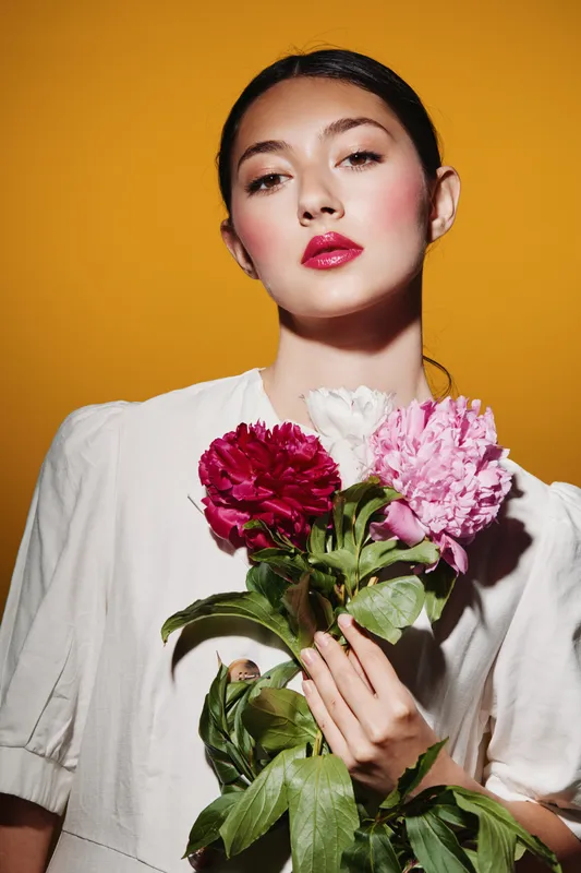 Fashion model with bold makeup holding red and pink flowers on a yellow background. Creative beauty concept focused on vivid colors and floral elements.