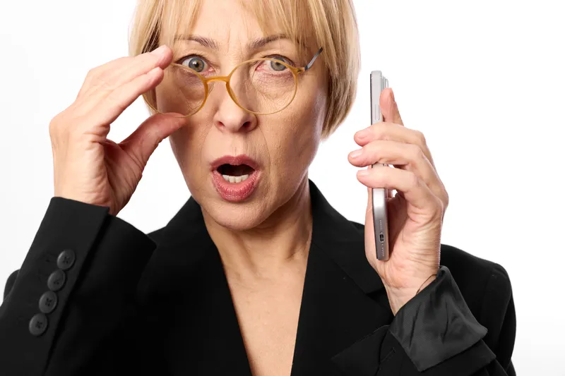 Senior woman with glasses holding a phone, surprised expression, hand touching frames, business suit, studio portrait background, communication moment