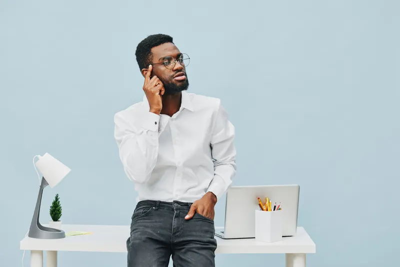 Thoughtful young man wearing white shirt standing at modern office desk with laptop, notepad, and stationery, pondering ideas and making decision in bright minimal workspace. People concept for work a