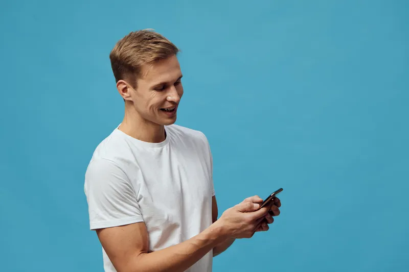 Smiling man wearing casual white t-shirt looking at mobile phone isolated on vibrant blue background studio portrait. People lifestyle concept