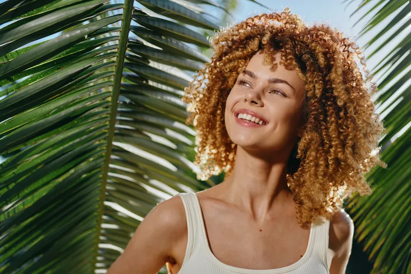 Smiling woman with curly hair enjoys sunlight outdoors against tropical green palm leaves, wearing casual white tank top in natural summer setting.