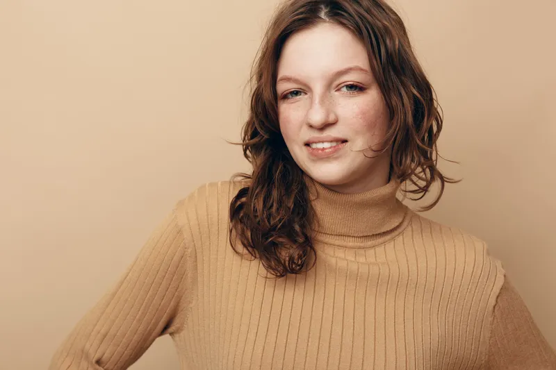 Young woman with curly brown hair wearing beige turtleneck sweater poses against beige background. Natural beauty portrait with soft lighting and casual style. Concept of confidence and simplicity.