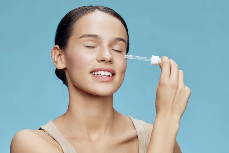 Young woman applying facial serum with a dropper, showing a joyful expression against a bright turquoise background Skincare, beauty routine, self care