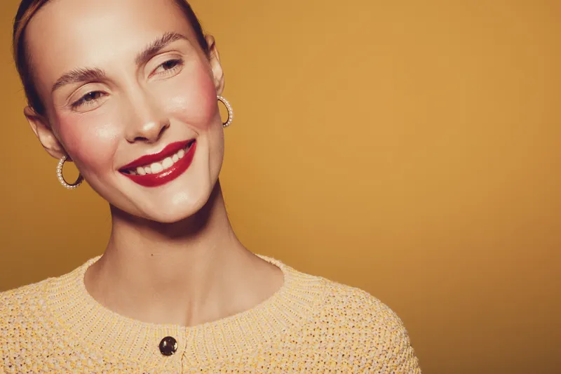 Smiling woman with red lipstick and hoop earrings looks away on yellow background. Close-up portrait with natural makeup and cozy sweater. Concept of happiness and beauty.