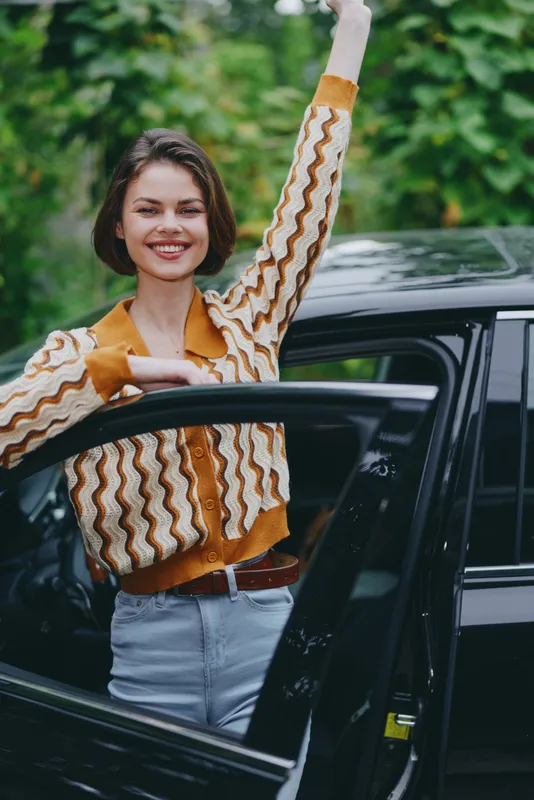A cheerful woman stands beside an open car door, raising a fist in celebration, wearing a retro knit sweater and jeans.