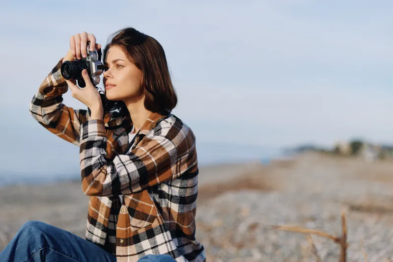 Woman photographer with camera on a beach, capturing sea and sky, outdoor scene in casual attire, focused moment by the shore.