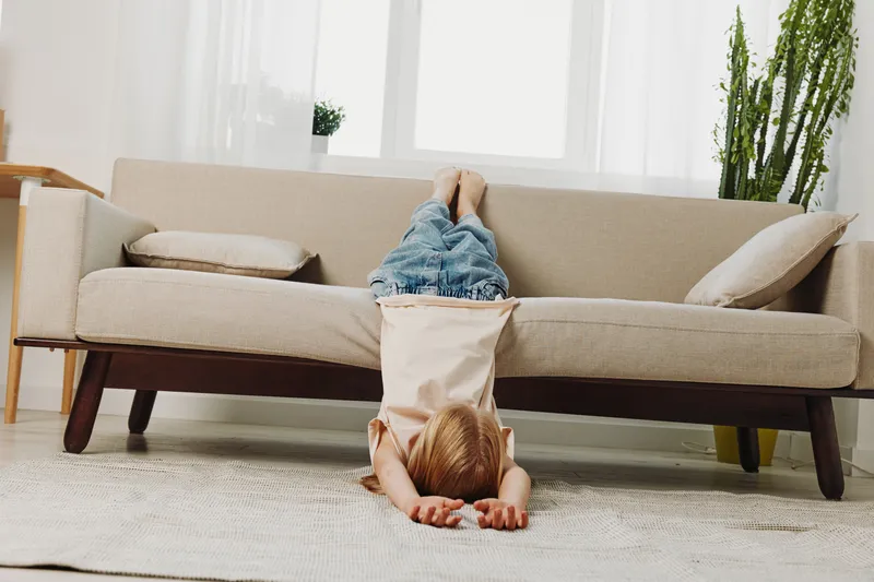 Young child lying upside down on floor with legs on sofa in bright living room. Cozy home interior with natural light, plants, and relaxed playful mood. Concept of childhood fun and comfort.
