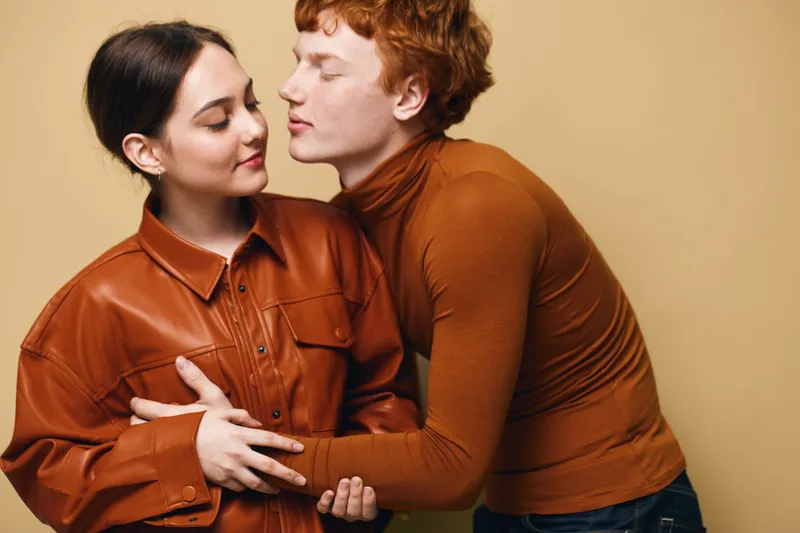 Young couple in warm brown clothing embracing tenderly against beige background. Woman with closed eyes and man leaning close showing affection and intimacy in studio portrait.