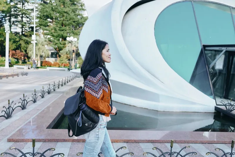 Woman walks along a modern outdoor plaza with a patterned sweater and backpack, passing sleek architecture and glass walls, conveying travel readiness, casual style, and urban exploration in daylight.