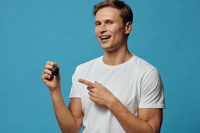 smiling young caucasian man hold car key fob in hand point finger wear white t-shirt blue background digital nomad concept