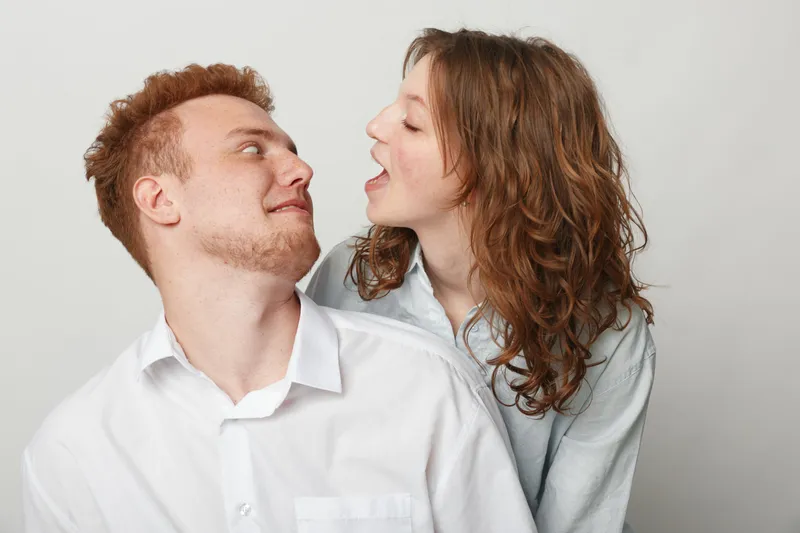 Young couple in casual white shirts interact playfully against a neutral background. The woman with curly hair leans toward the man with red hair, creating a lively and intimate moment.