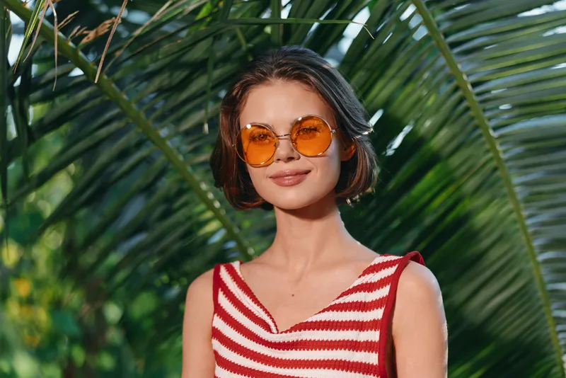 A stylish woman stands outdoors amid lush palm leaves, wearing a striped red and white top and round orange sunglasses. She smiles softly, exuding sunny confidence and relaxed charm in a vibrant