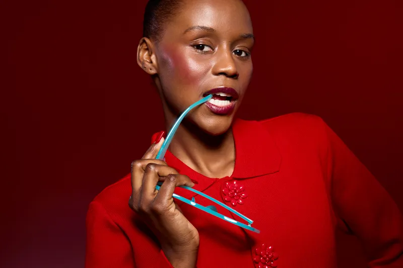 Portrait of elegant dark skinned African woman in red sweater holding blue glasses near lips on red background. Concept for fashion, beauty, and confident female model with glossy makeup.