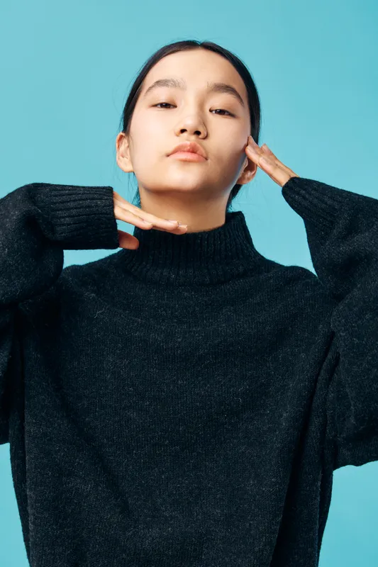 Young woman posing confidently with hands near face wearing black sweater against blue background. Portrait, casual style, fashion concept with natural expression and minimal makeup.