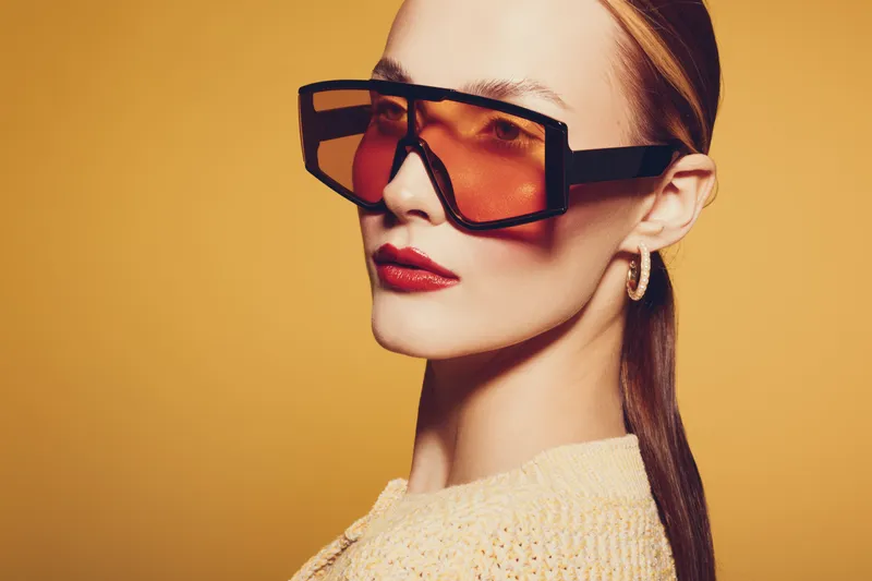 Stylish woman with oversized orange sunglasses and red lipstick poses against yellow background. Fashion portrait with modern accessories and confident expression.
