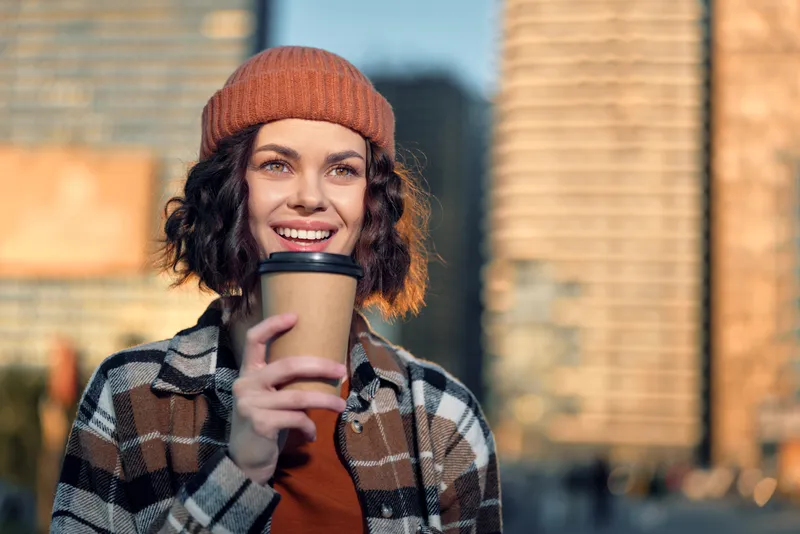 Young woman in beanie holding coffee with a warm smile in an urban street at golden hour, candid lifestyle portrait with authenticity, golden hour glow, mindful living and emotional storytelling.
