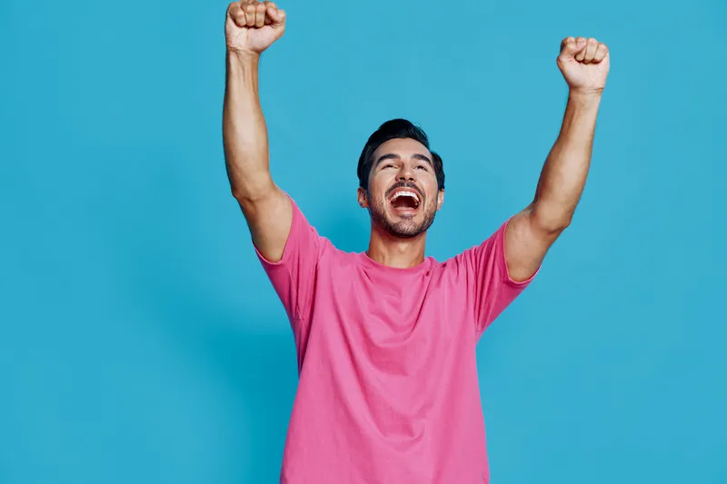 Full body ecstatic happy young man wearing pink t-shirt celebrates with arms raised against a solid blue background in a joyful studio setting. Lifestyle concept.