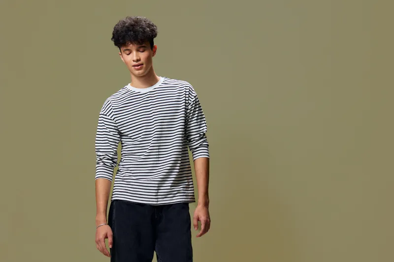 young man wearing striped long sleeve shirt standing with relaxed posture against plain olive background, casual style, thoughtful expression