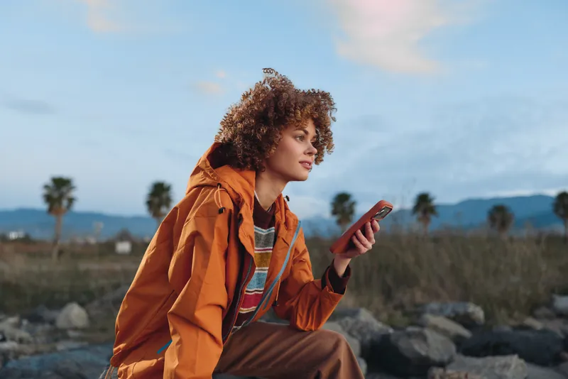 Woman lifestyle outdoors with curly hair wearing orange jacket holding smartphone by a rocky coast, looking relaxed, blue sky and distant mountains, warm travel mood, natural sunset light